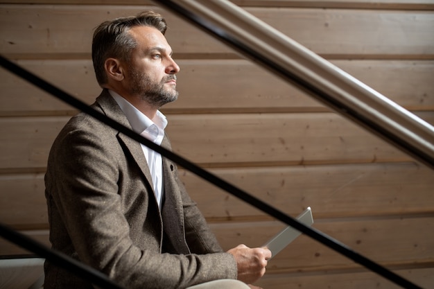 Nachdenklicher Geschäftsmann mit Touchpad, der auf Treppe durch Holzwand innerhalb des zeitgenössischen Restaurants oder des Geschäftszentrums sitzt