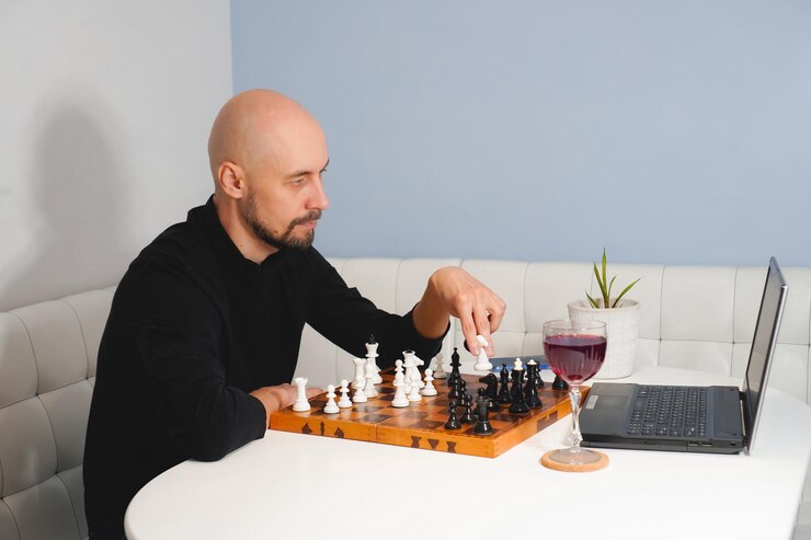 Nachdenklicher glatzköpfiger Mann mit Bart spielt Schach an Bord in der Nähe von Laptop und Weinglas