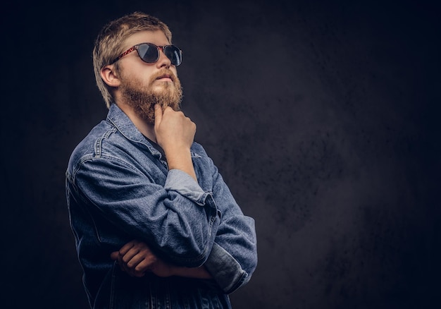 Nachdenklicher Hipster-Typ mit Sonnenbrille in Jeansjacke, der mit der Hand am Kinn posiert. Getrennt auf einem dunklen Hintergrund.