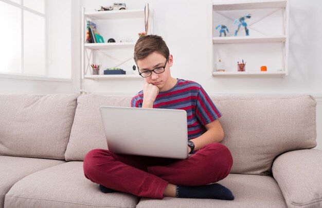 Nachdenklicher Junge mit Laptop-Computer auf dem Sofa zu Hause. Kind macht Hausaufgaben, sucht nach Informationen im Internet, tippt auf der Tastatur