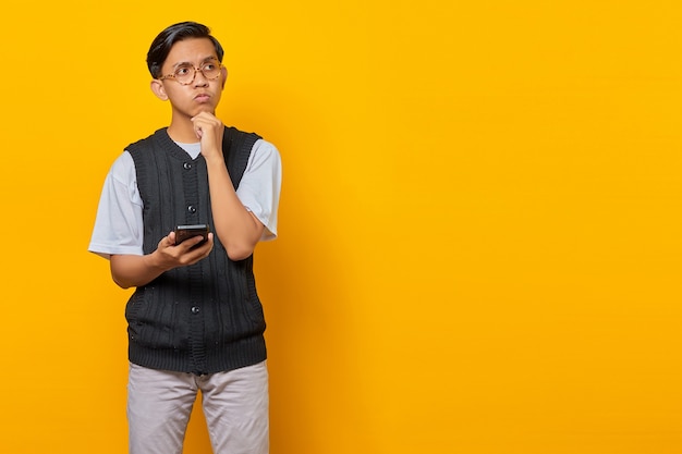 Nachdenklicher junger asiatischer Mann, der Handy mit den Fingern am Kinn benutzt und beiseite schaut