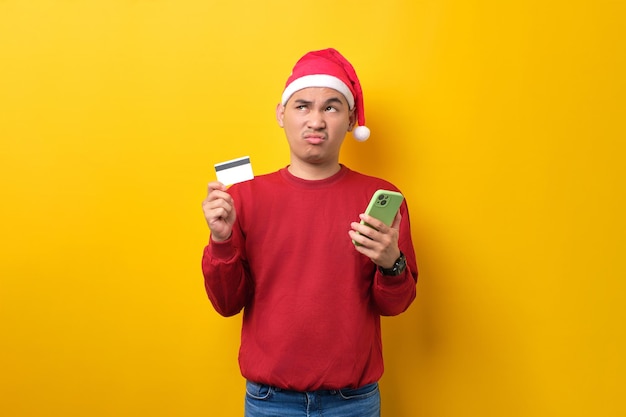 Nachdenklicher junger asiatischer Mann in Weihnachtsmütze mit Handy und Kreditkarte, der nach oben schaut und über gelbe Studiohintergrundfeier Weihnachtsferien und Neujahrskonzept nachdenkt