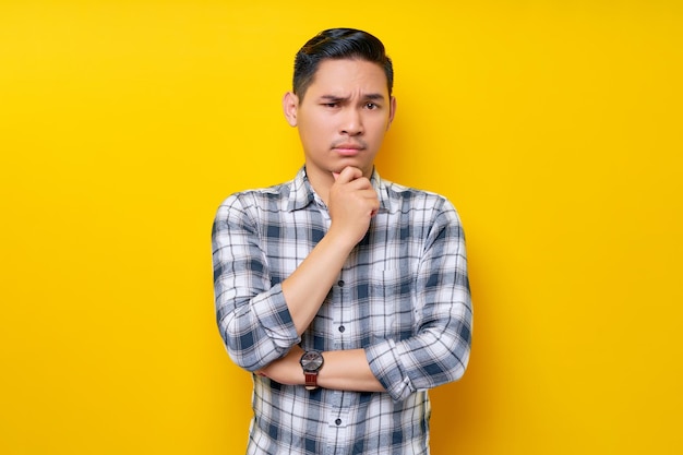 Nachdenklicher junger gutaussehender asiatischer Mann in legerer Kleidung hält seine Hand auf seinem Kinn isoliert auf gelbem Hintergrund Lifestyle-Konzept für Menschen
