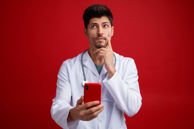 Nachdenklicher junger männlicher Arzt, der eine medizinische Uniform und ein Stethoskop um den Hals trägt und ein Handy hält, das die Hand am Kinn hält und isoliert auf rotem Hintergrund nach oben schaut
