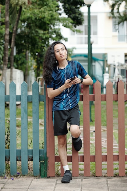 Nachdenklicher junger Mann in Sportkleidung, der sich nach dem Morgenlauf auf einen Zaun stützt und Musik in Ohrhörern hört