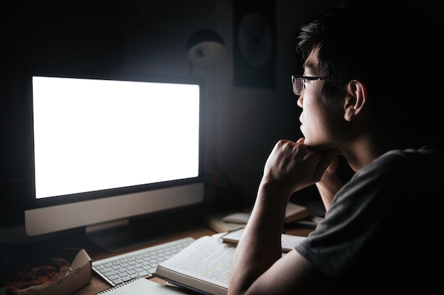 Nachdenklicher junger Mann mit Brille, der mit einem Computer mit leerem Bildschirm in einem dunklen Raum studiert