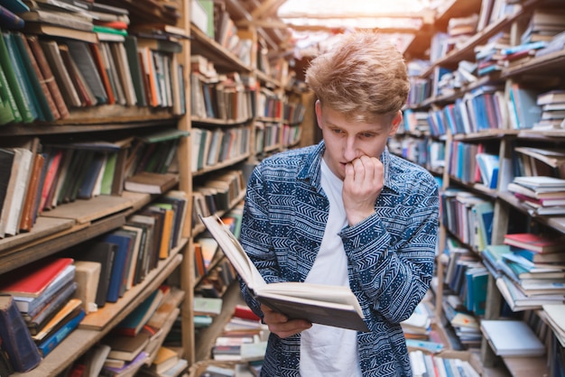 Nachdenklicher junger Mann steht in einer gemütlichen Bibliothek, liest ein Buch und beißt sich auf die Nägel.