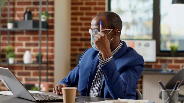 Nachdenklicher männlicher Berater, der über Finanzstrategie nachdenkt, um Geschäftsstatistiken auf dem Laptop zu erstellen. Nachdenklicher Manager mit Gesichtsmaske Brainstorming-Ideen, sich unsicher fühlen und Entscheidungen erwägen.