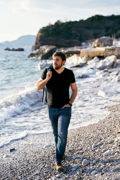 Nachdenklicher Mann, der auf einem Kiesstrand mit einer Jeansjacke über der Schulter vor dem Hintergrund spaziert