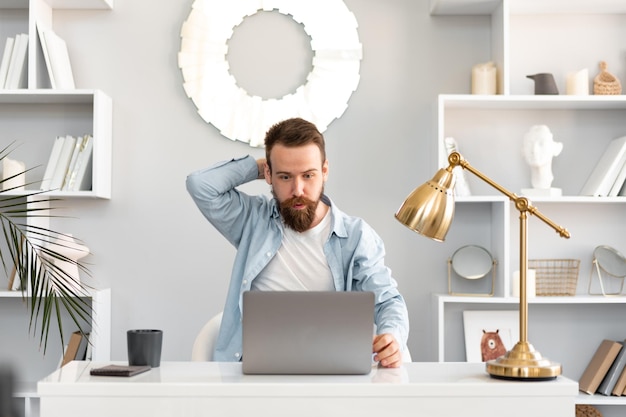 Nachdenklicher müder junger bärtiger Mann, der am Tisch sitzt und am Laptop arbeitet