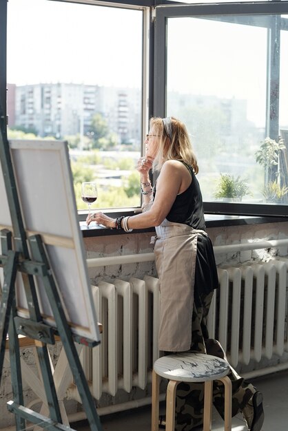 Nachdenklicher reifer Künstler im Vorfeld, der das Stadtbild durch das Fenster betrachtet und Inspiration im eigenen Studio findet