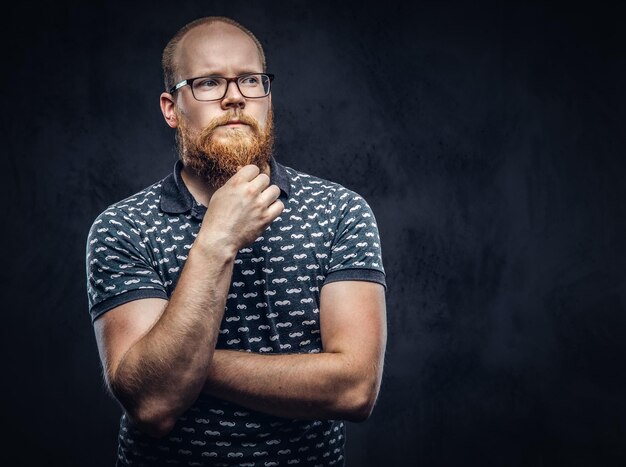 Nachdenklicher rothaariger bärtiger Mann mit Brille, gekleidet in ein T-Shirt, das mit einer Hand am Kinn posiert. Getrennt auf einem dunklen strukturierten Hintergrund.