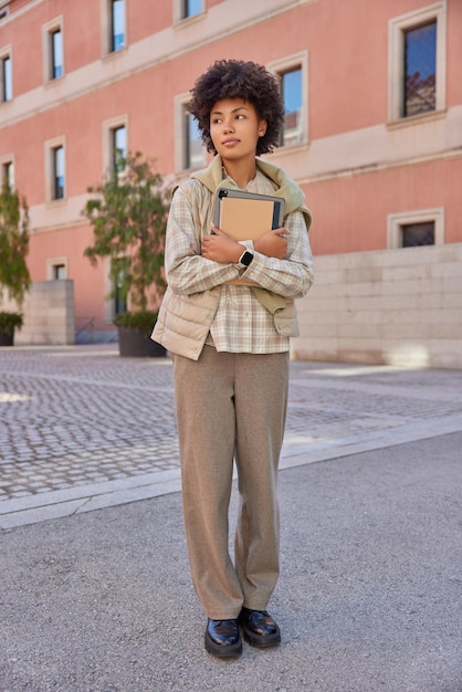 Nachdenklicher Student hält modernes digitales Tablet und Notebook trägt stilvolle Kleidung Spaziergänge im Freien gegen Gebäude