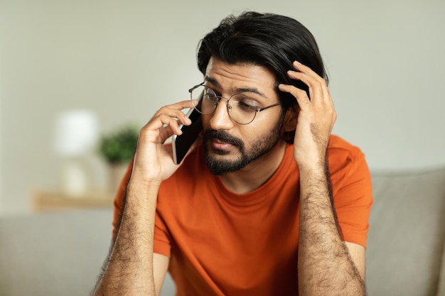 Nachdenklicher, tausendjähriger Mann aus dem Nahen Osten mit Brille telefoniert im Wohnzimmer