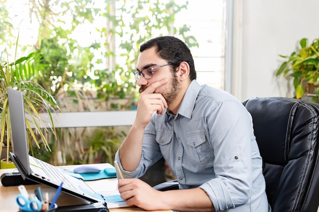 Nachdenklicher Unternehmer, der sich Notizen macht, während er im Home Office am Laptop arbeitet