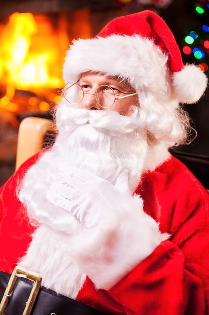 Nachdenklicher Weihnachtsmann. Nachdenklicher Weihnachtsmann sitzt an seinem Stuhl und berührt Bart mit Kamin und Weihnachtsbaum im Hintergrund