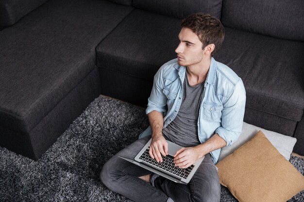 Nachdenklicher zufälliger Mann, der am Laptop arbeitet und wegschaut, während er zu Hause auf dem Teppich sitzt