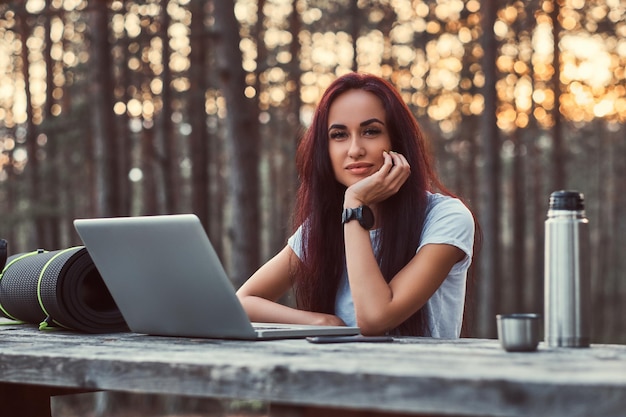 Nachdenkliches Hipster-Mädchen in weißem Hemd, das in die Kamera schaut, während es auf einer Holzbank mit offenem Laptop in einem schönen Herbstwald sitzt.