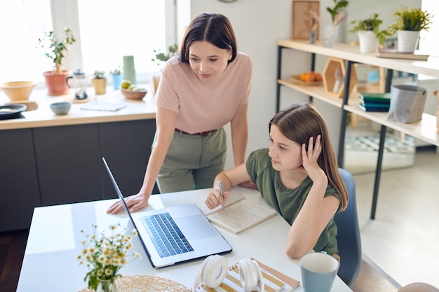 Nachdenkliches junges Mädchen, das Notizen im Arbeitsbuch macht, während Online-Vortrag auf Laptop unter Kontrolle der Mutter beobachtet