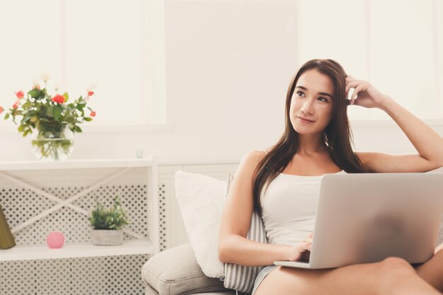 Nachdenkliches lächelndes Mädchen, das mit Laptop bequem auf beigem Sofa sitzt und online SMS schreibt. Platz kopieren