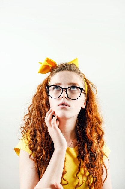 Nachdenkliches lockiges rothaariges Mädchen mit Brille mit einer gelben Schleife auf dem Kopf, das ein gelbes T-Shirt trägt und nach oben schaut und nachdenkt