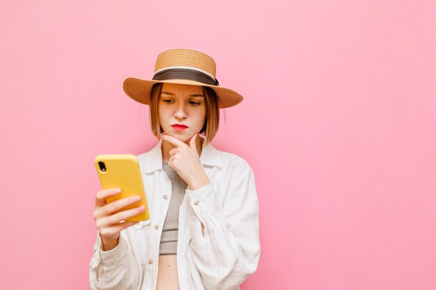 Nachdenkliches Mädchen mit Hut und Hemd an der Spitze steht auf rosa Hintergrund mit Smartphone in der Hand