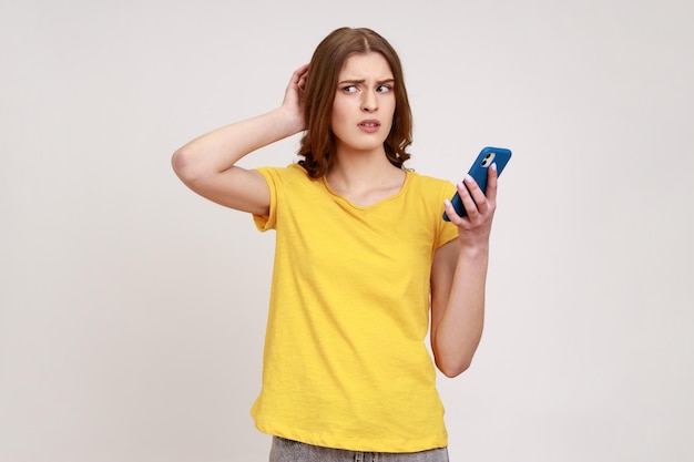 Nachdenkliches, nachdenkliches Teenager-Mädchen im gelben, lässigen Stil T-Shirt, das mit dem Handy steht, das den Kopf berührt und Zweifel im Verdacht hat, Studioaufnahme isoliert auf grauem Hintergrund