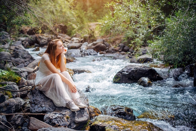 Nachdenkliches romantisches junges Mädchen in weißem Kleid, das am Ufer des Gebirgsflusses zwischen grünen Bäumen sitzt Schöne langhaarige Frau entspannt sich allein mit der Natur Harmony Happy Lifestyle-Konzept