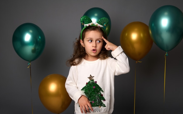 Nachdenkliches süßes Mädchen legt ihren Finger auf ihre Schläfe und schaut nachdenklich nach unten, posiert vor grauem Hintergrund mit schönen goldenen und glänzend grünen metallischen Luftballons mit Kopienraum für Weihnachtswerbung