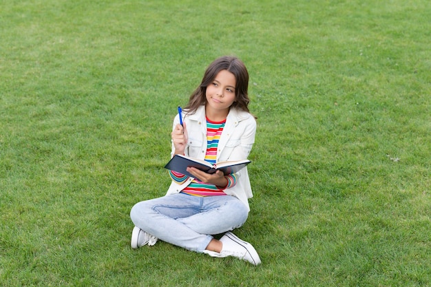 Nachdenkliches Teenager-Kind, das sich Notizen im Notizbuch macht, das auf Gras sitzt und sich Notizen macht