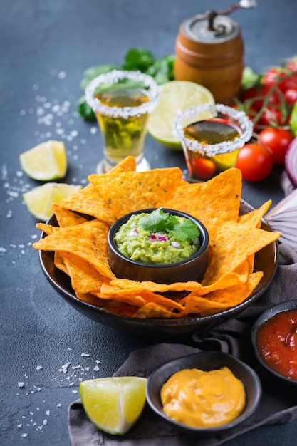 Nachos de chips de tortilla de maíz amarillo con guacamole, salsa de chile jalapeño candente y salsa de queso con tequila sobre una mesa oscura.