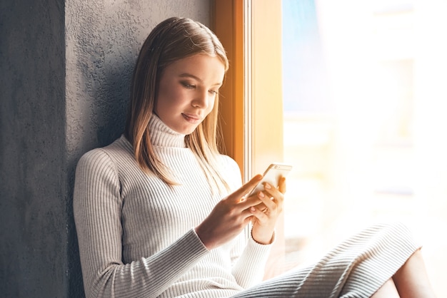 Nachricht an ihn. Schöne junge Frau, die ihr Smartphone benutzt, während sie zu Hause auf der Fensterbank sitzt