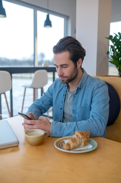 Nachricht. Konzentrierter bärtiger junger Mann, der aufmerksam in das Smartphone blickt, das tagsüber mit Laptop und Kaffee im Café am Tisch sitzt