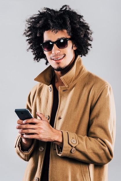 Nachricht von einem Freund. Fröhlicher junger Afrikaner mit Sonnenbrille, der mit einem Lächeln in die Kamera schaut und das Smartphone im Stehen hält