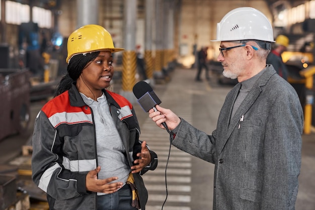 Nachrichtenreporter im Arbeitshelm, der das Mikrofon vor der Arbeiterin hält und sie während ihrer Arbeit in der Fabrik interviewt