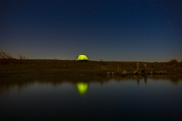 Nachthimmel mit Sternen über dem Touristenzelt am Fluss. Die Landschaft wurde mit einer langen Belichtungszeit fotografiert.