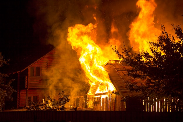 Nachts brennendes Holzhaus. Leuchtend orange Flammen und dichter Rauch unter dem Ziegeldach auf dunklem Himmel, Baumsilhouetten und Wohnnachbarhäuschenhintergrund. Katastrophen- und Gefahrenkonzept.