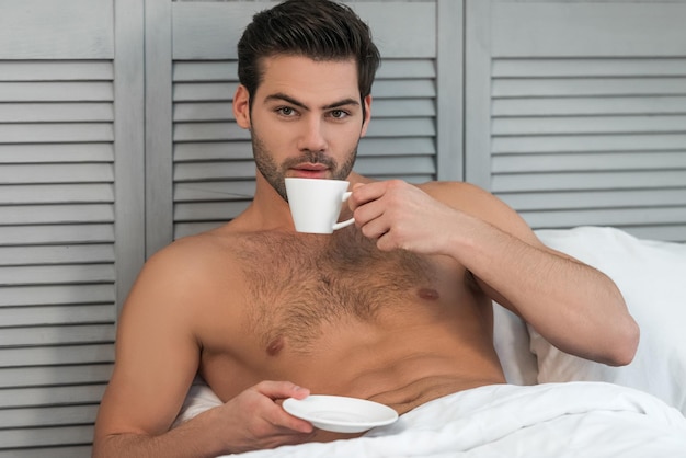 Nackter gutaussehender Mann, der im Bett Kaffee trinkt und in die Kamera schaut