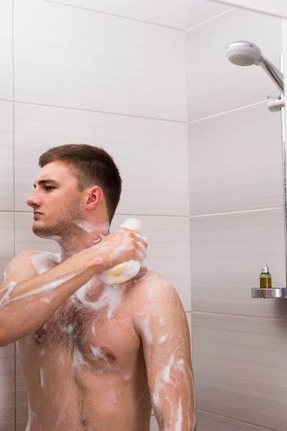 Nackter junger Mann reibt sich ein Schaumbad, während er in der Duschkabine mit transparenten Glastüren im Badezimmer steht
