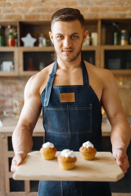 Nackter Mann in der Schürze hält Tablett mit süßem Dessert auf der Küche. Nackte männliche Person, die Frühstück zu Hause vorbereitet, Essenszubereitung ohne Kleidung