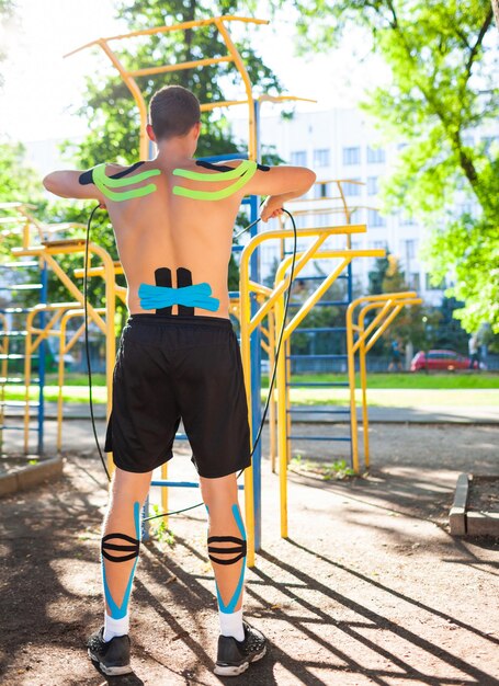 Nackter muskulöser Mann trainiert mit Fitnessseil am Sportplatz. Rückansicht eines jungen, nicht erkennbaren Bodybuilders mit elastischer Kinesiologie, die beim Körpertraining im Freien aufnimmt. Rehabilitationskonzept.