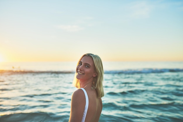 Nada como el aire fresco del océano Retrato recortado de una atractiva mujer joven sonriendo sobre su hombro en la playa al atardecer