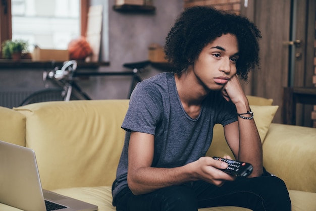 Nada de interessante para assistir. Jovem africano segurando o controle remoto e parecendo entediado enquanto assiste TV no sofá de casa