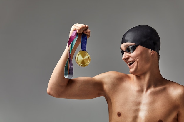 El nadador con una medalla sobre un fondo gris se regocija en la victoria, un atleta en excelente forma física celebra una victoria sosteniendo una medalla en la mano, concepto de victoria, fondo gris, espacio de copia