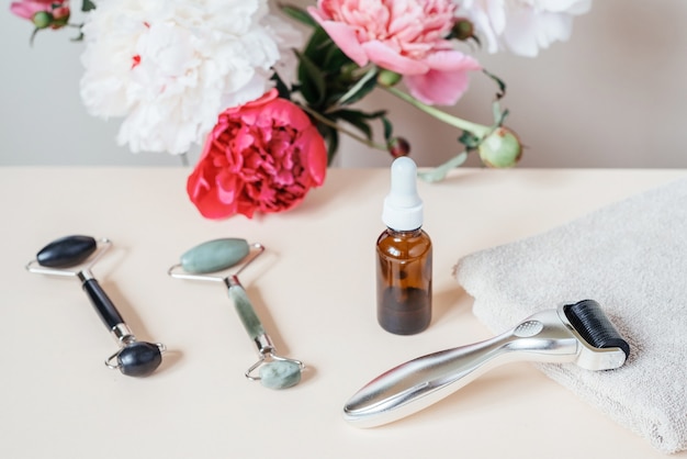 Nadel-Derma-Roller, Jade-Guasha-Massagerollen und Serumflasche mit Pfingstrosenblüten im Hintergrund, Mikronadeln für zu Hause und Hautpflege