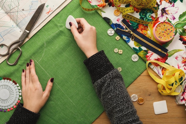 Nähwerkstatt oder Modedesigner bei der Arbeit. Draufsicht auf weibliche Hände, die Tuchquadrat mit Seifenstück markieren. Unordentlicher Tisch mit Schneiderzubehör und Mustern, Kopierraum