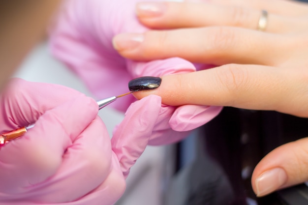 Nagelpflege und Maniküre-Konzept. Nahaufnahme-Maniküristhände in den rosa Handschuhen malt schwarzen Nagellack auf den Nägeln des Kunden.