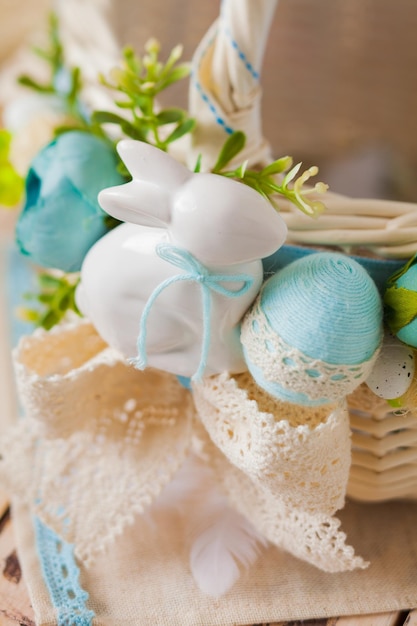 Nahansicht des schönen weißen Keramikkaninchens, das am Griff des Korbs befestigt ist, zusammen mit blauem Ei, das mit Baumwollfäden umwickelt ist, und beiger Spitze Osterkonzept