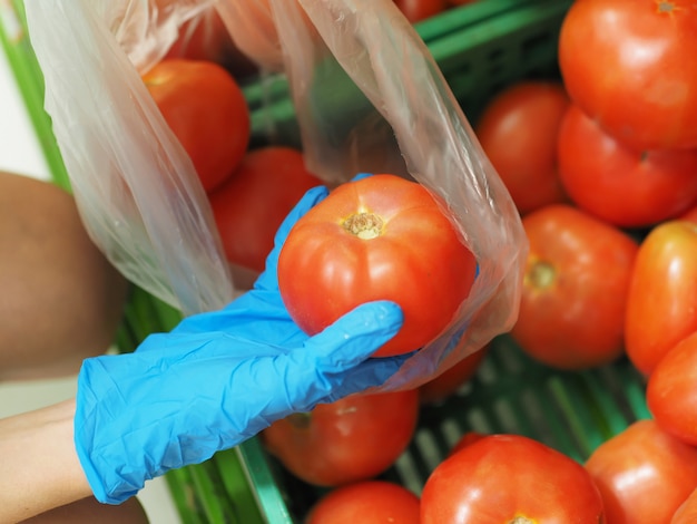 Nahansicht. Weibliche Hände in blauen Handschuhen, die eine Tomate im Supermarkt während des pandemischen Coronavirus COVID-19 wählen.