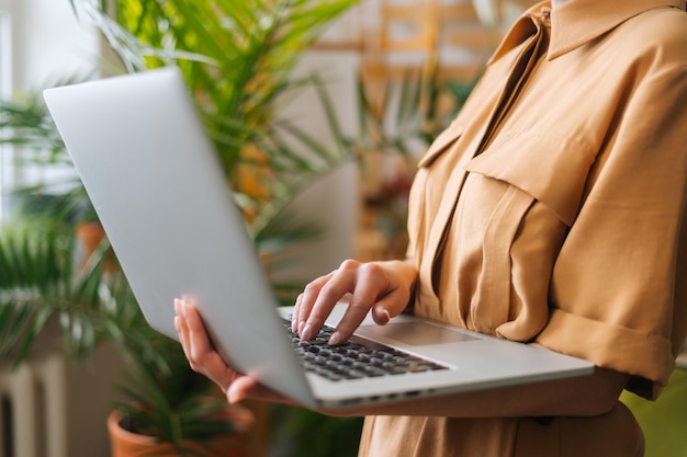 Nahaufnahme, abgeschnittene Aufnahme einer nicht erkennbaren Geschäftsfrau, der Besitzerin eines Blumenladens, die mit einem Laptop steht, der in der Hand zwischen grünen Pflanzen hält